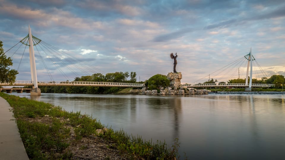 A suspension bridge over a calm river, with a statue on a rock in the center.