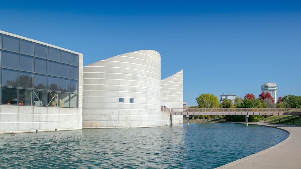 A modern building with a curved facade and a glass section, a bridge over a body of water, and a tower in the background.
