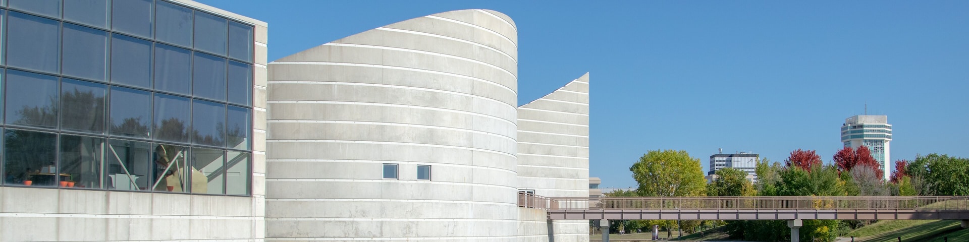A modern building with a curved facade and a glass section, a bridge over a body of water, and a tower in the background.