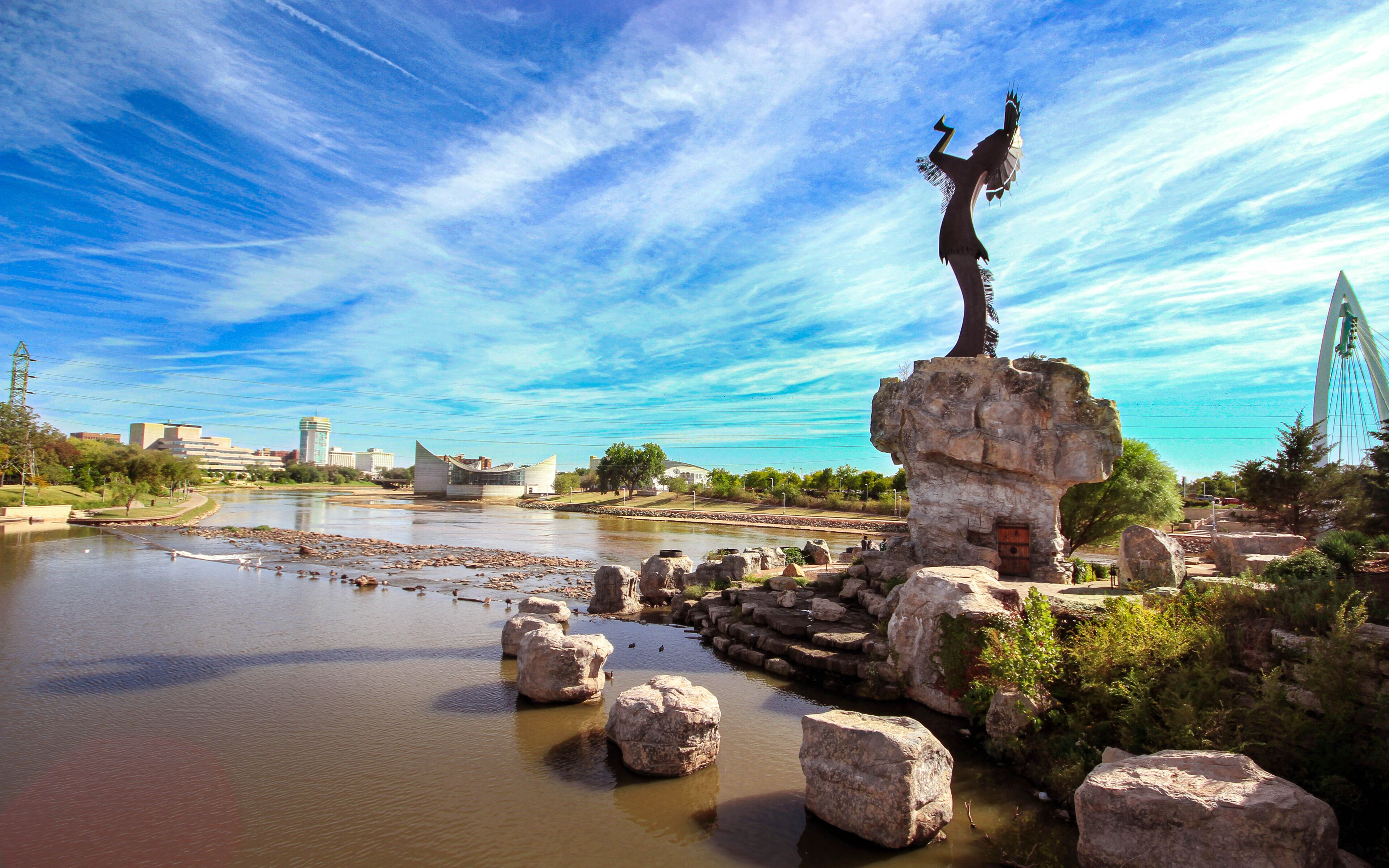 A river with a sculpture on a rock, a bridge, and buildings in the background.