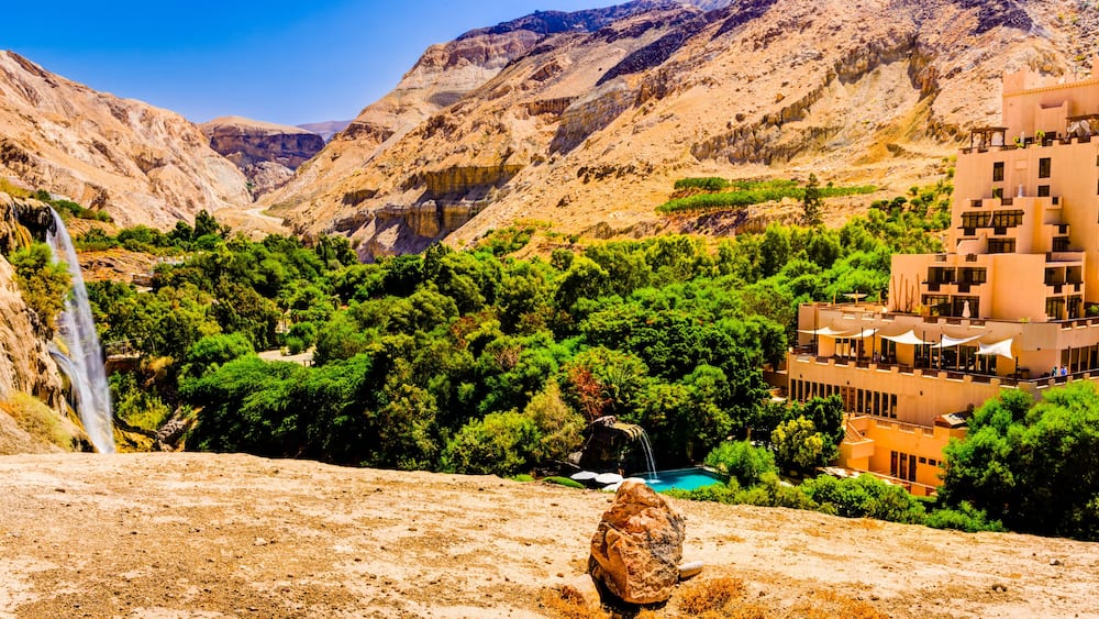 A mountainous landscape with a waterfall, lush greenery, and a modern building complex.