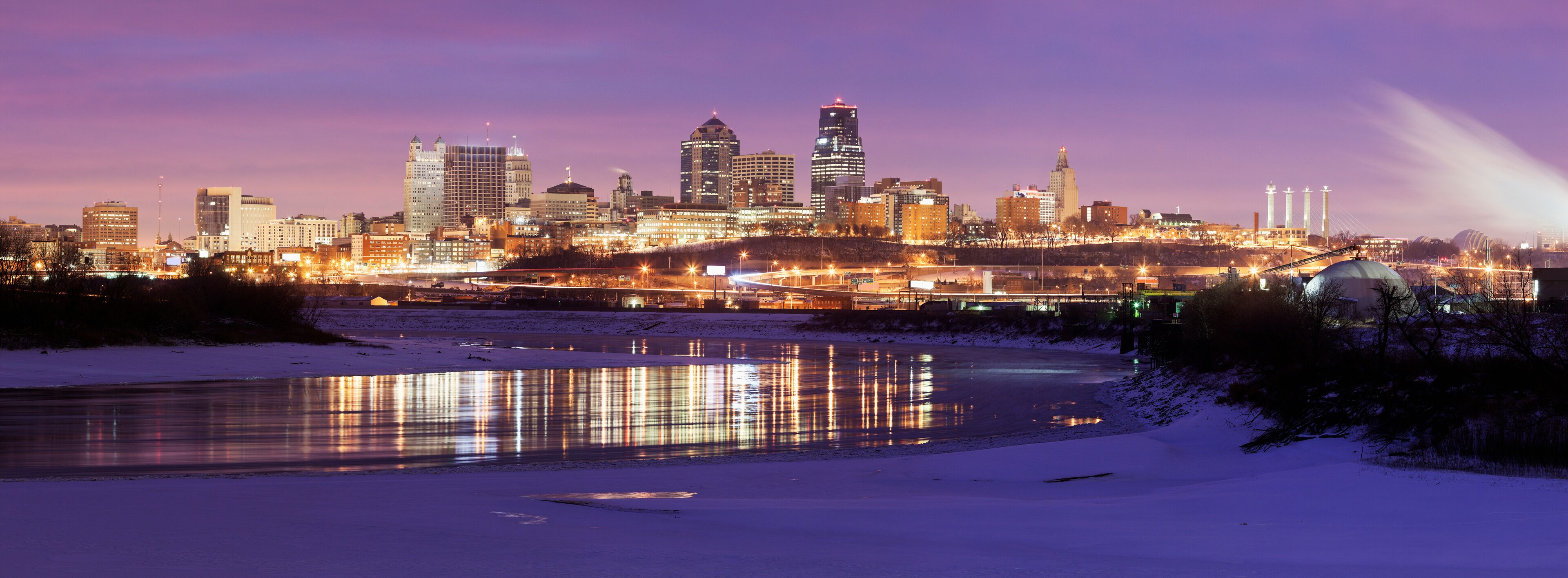 A city skyline at dusk with illuminated buildings and a frozen river reflecting lights.
