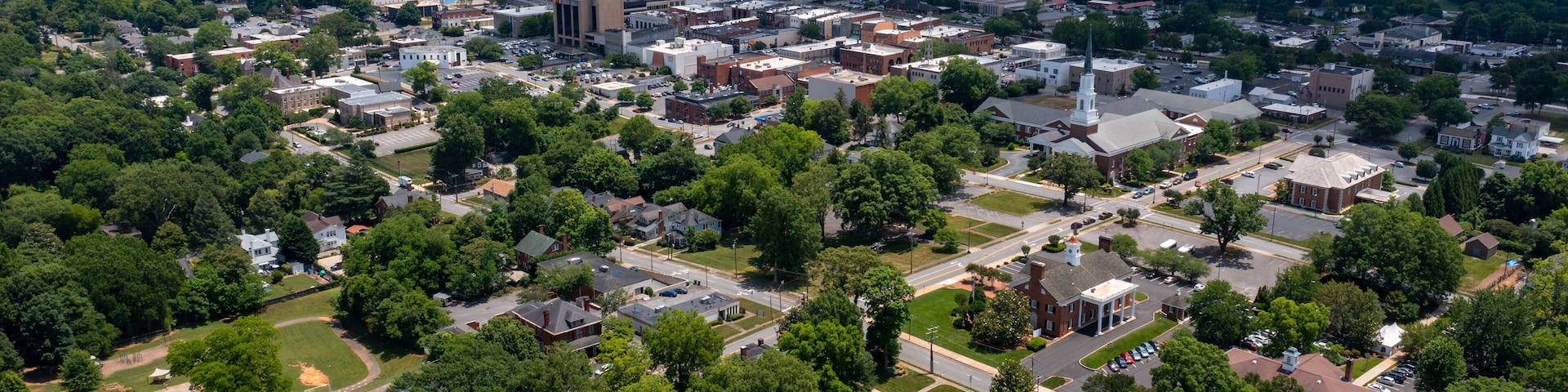 An aerial view of a town with a mix of residential and commercial buildings, surrounded by greenery.