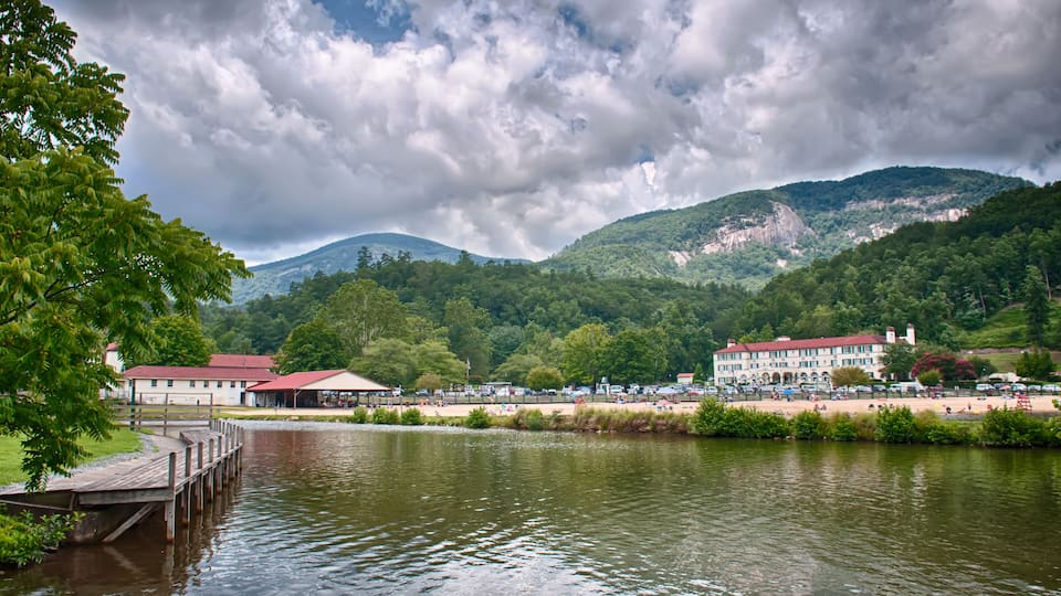 A lake with a wooden dock, buildings, and a mountainous backdrop under a cloudy sky.