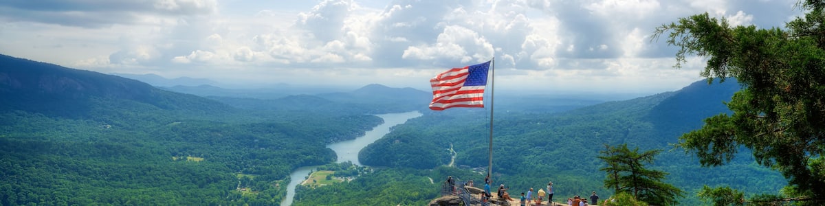 A scenic overlook with a flag on a rock formation, a river in the valley below, and lush greenery.