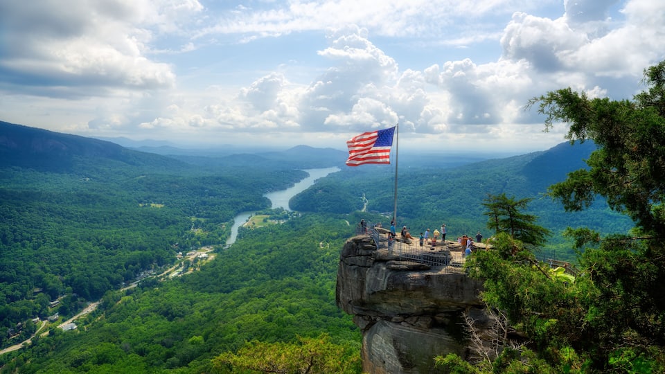 A scenic overlook with a flag on a rock formation, a river in the valley below, and lush greenery.