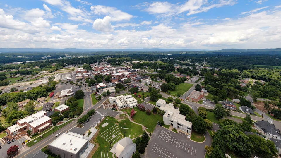 A town with a mix of commercial and residential buildings, surrounded by greenery and a clear sky.