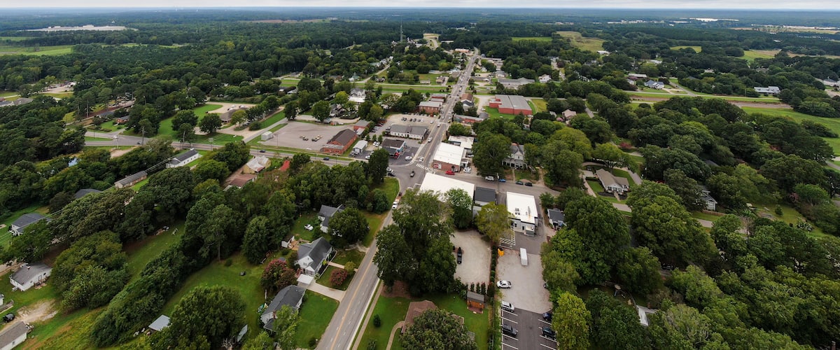 An aerial view of a town with a central road, surrounding greenery, and various buildings.