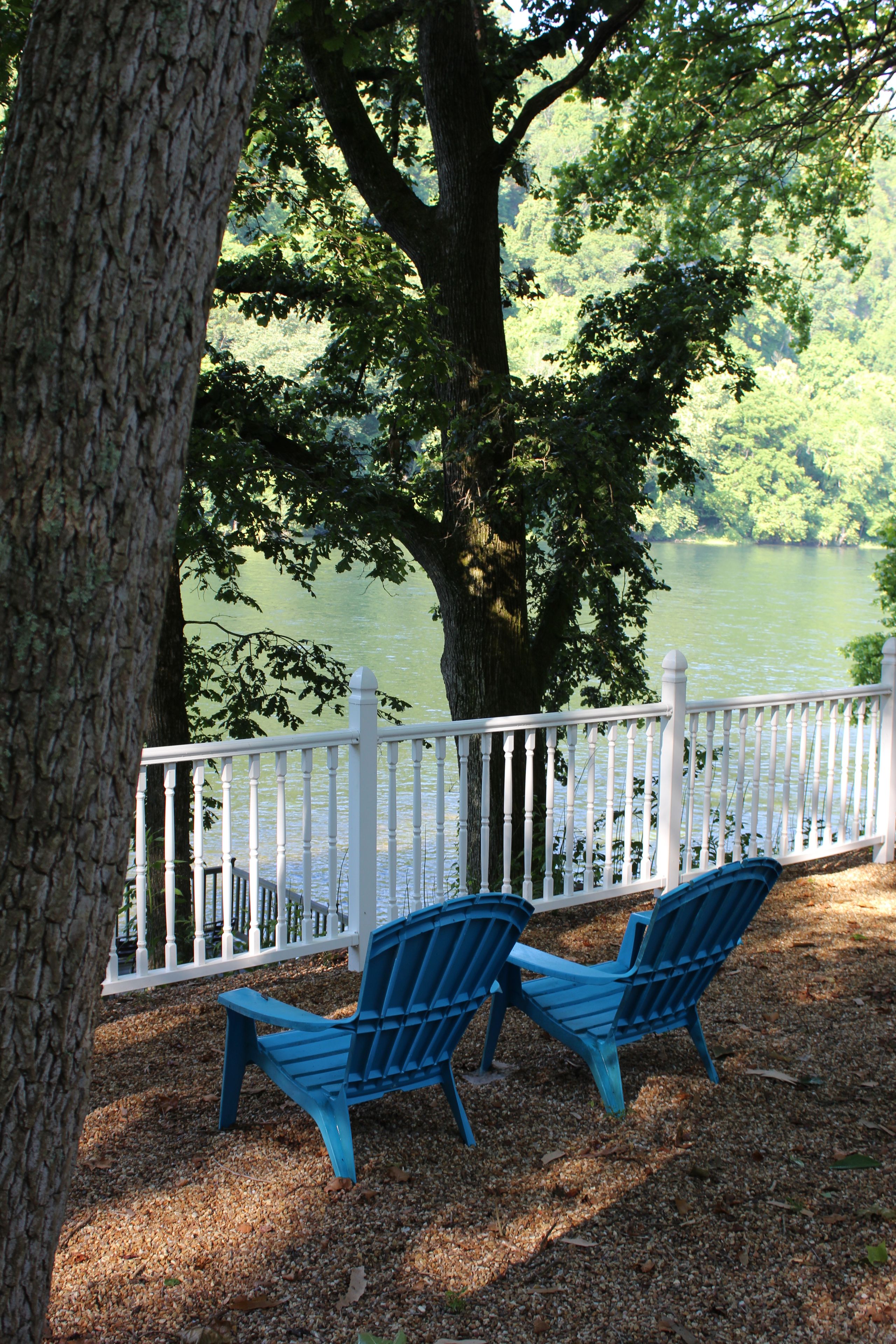 A lakeside view with a white fence and two blue Adirondack chairs.
