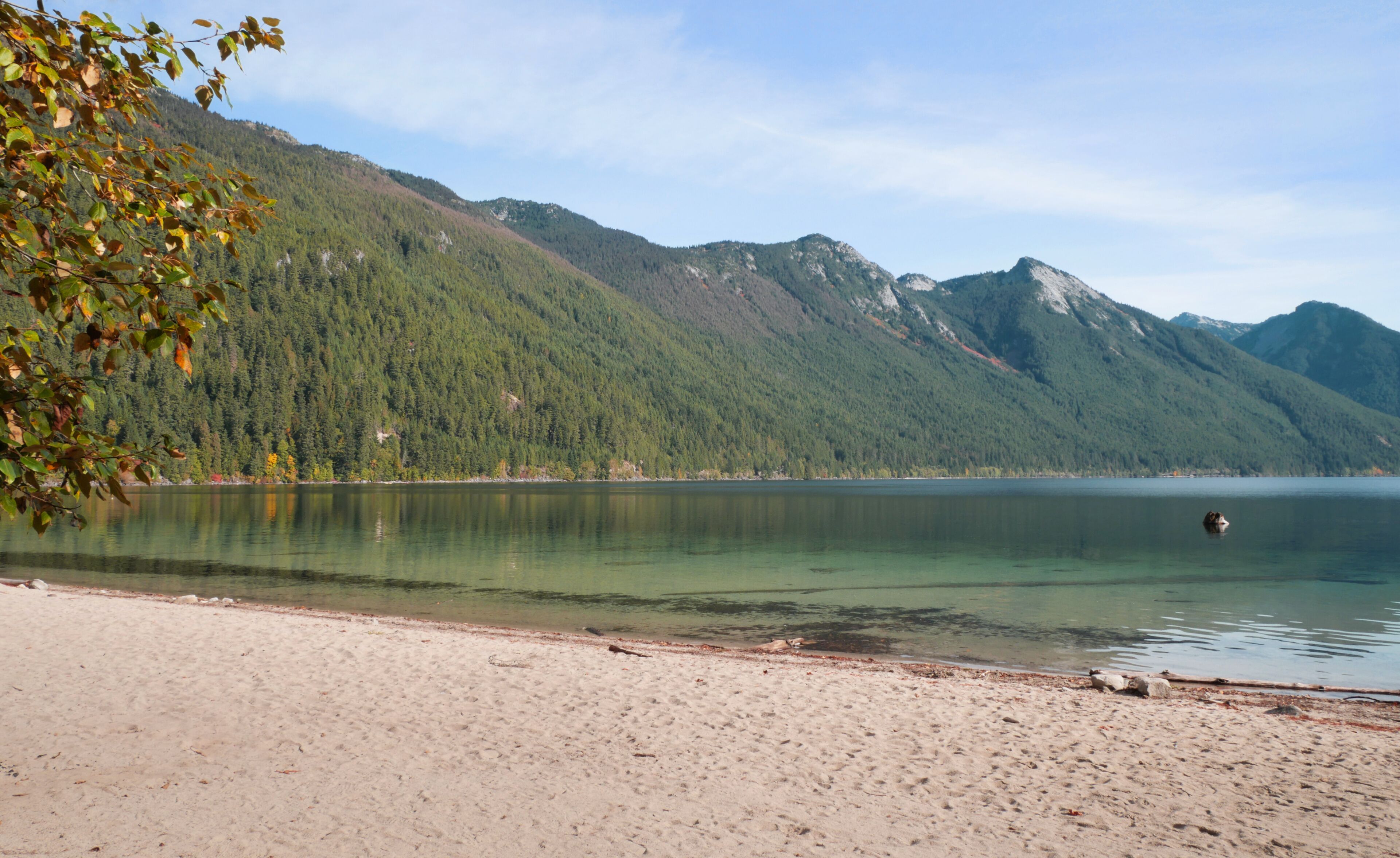 A lake with a sandy beach, surrounded by forested mountains.