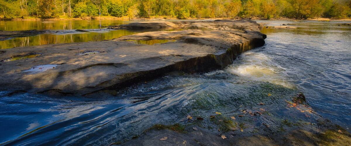 A river with a large rock formation in the middle, surrounded by trees and a clear sky.
