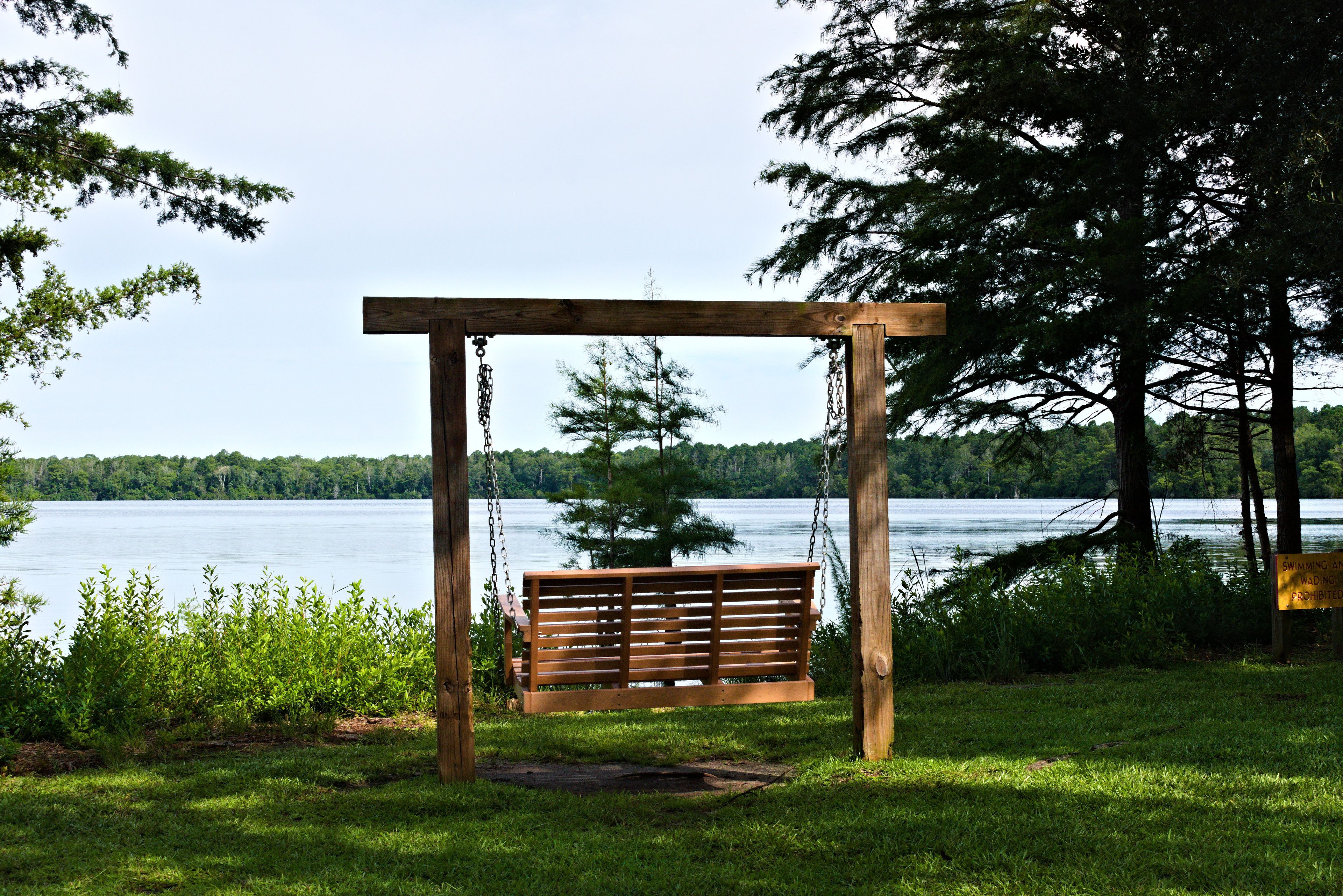 A wooden swing set with a bench, overlooking a calm lake surrounded by trees.