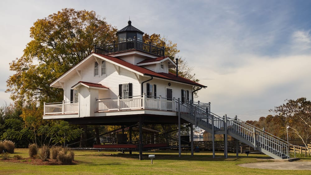 A white house with a balcony and a tower, elevated on stilts, surrounded by trees and a grassy area.
