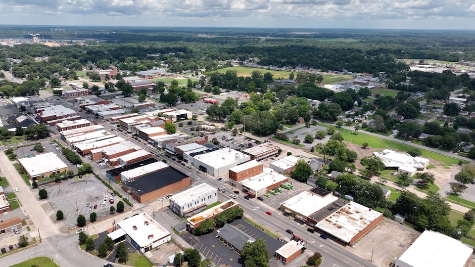 An aerial view of a town with a mix of commercial buildings, a large parking lot, and surrounding greenery.