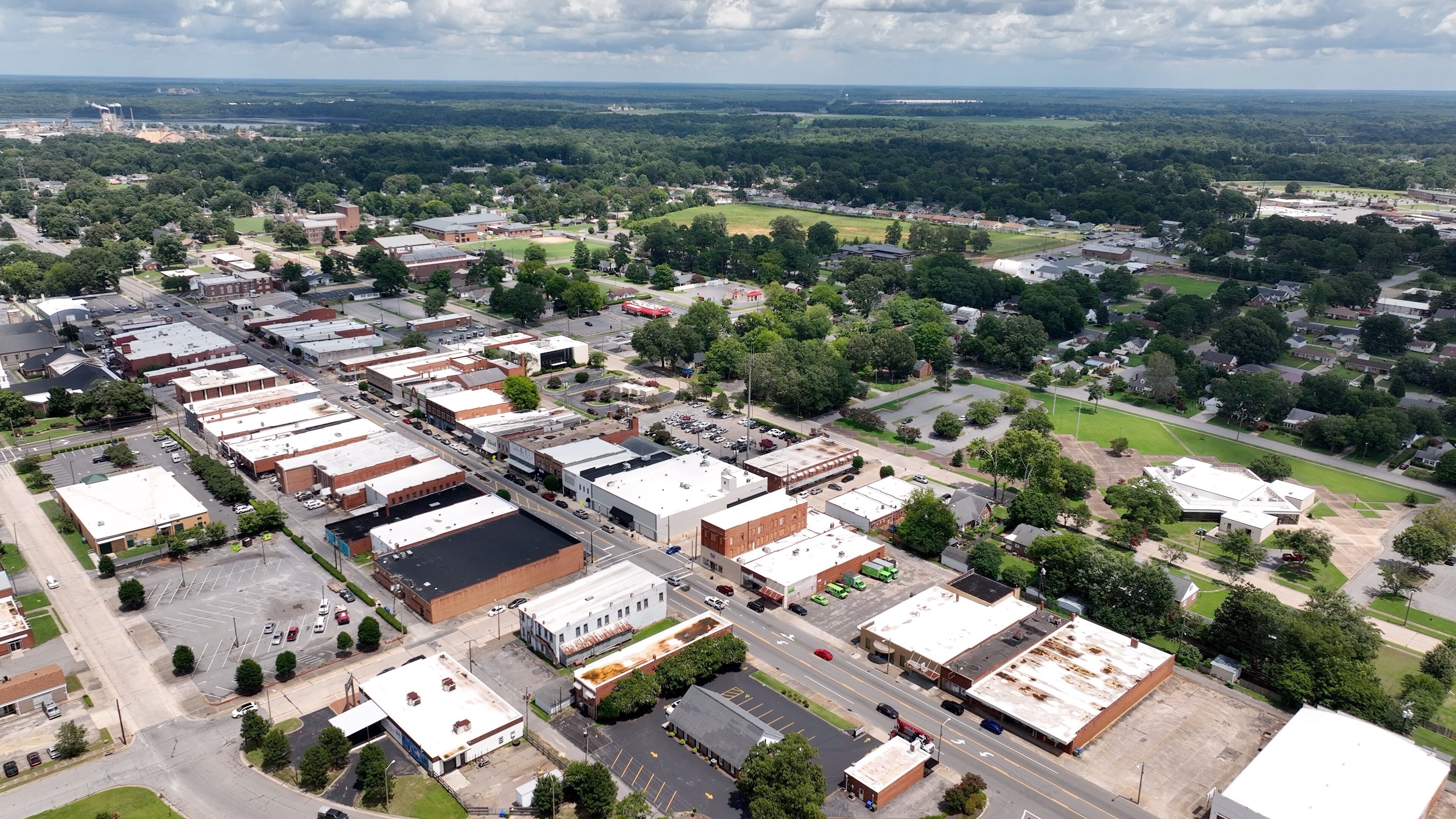 An aerial view of a town with a mix of commercial buildings, a large parking lot, and surrounding greenery.