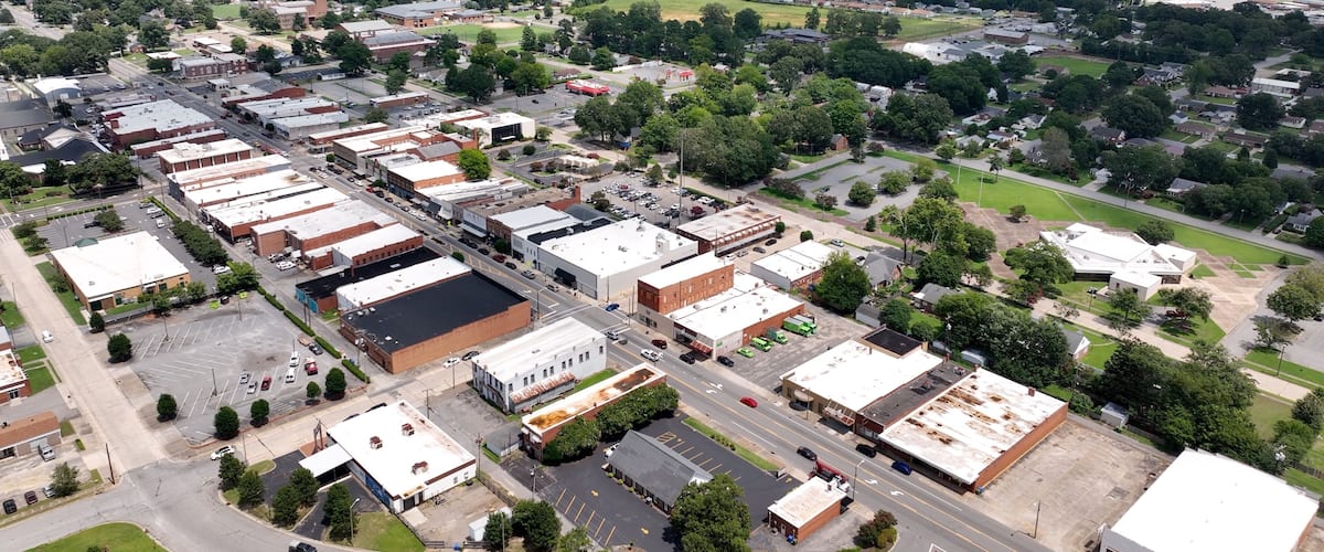 An aerial view of a town with a mix of commercial buildings, a large parking lot, and surrounding greenery.