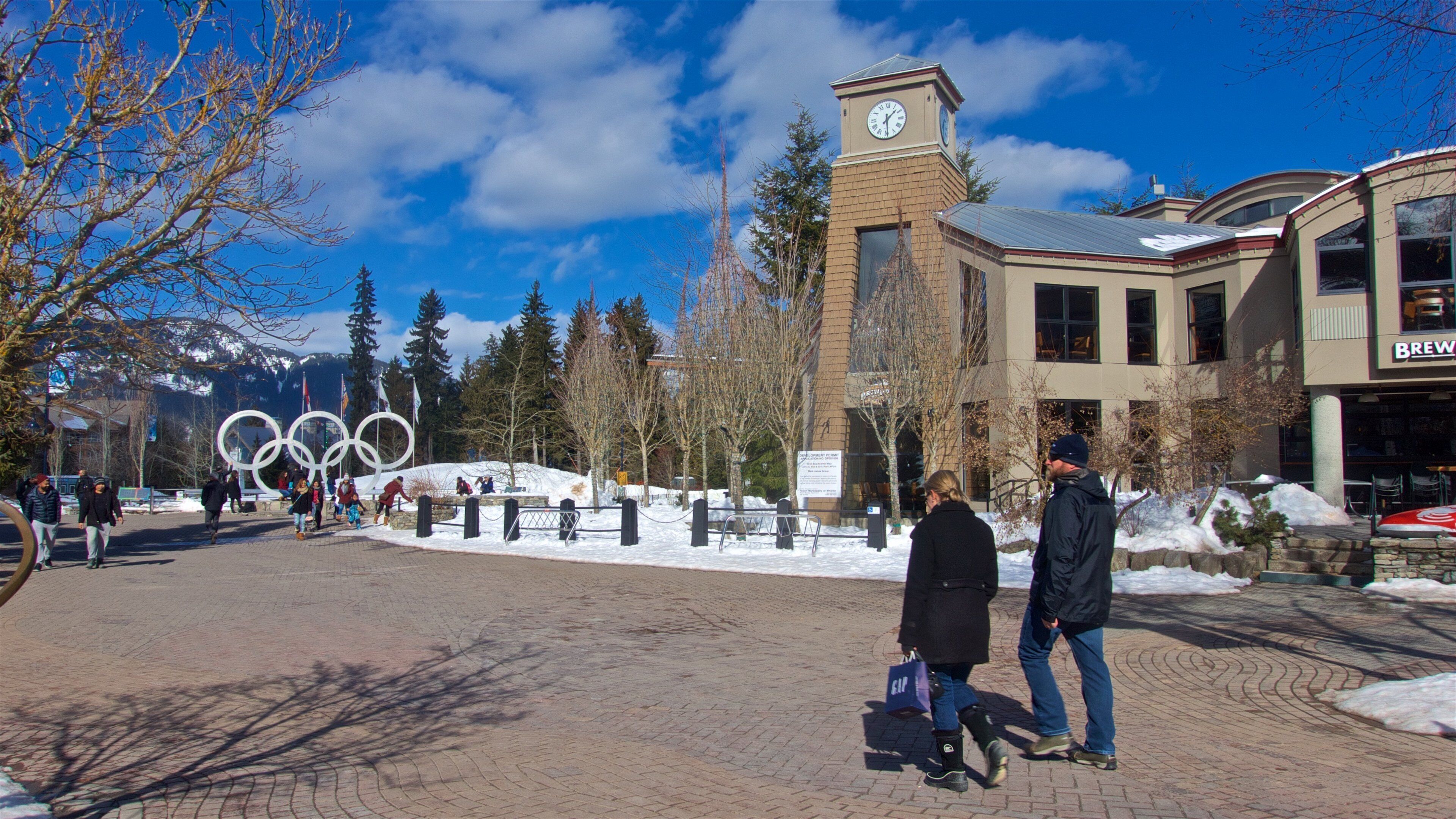 A snowy plaza with the Olympic rings sculpture, a clock tower, and a building with a 'Brewery' sign.