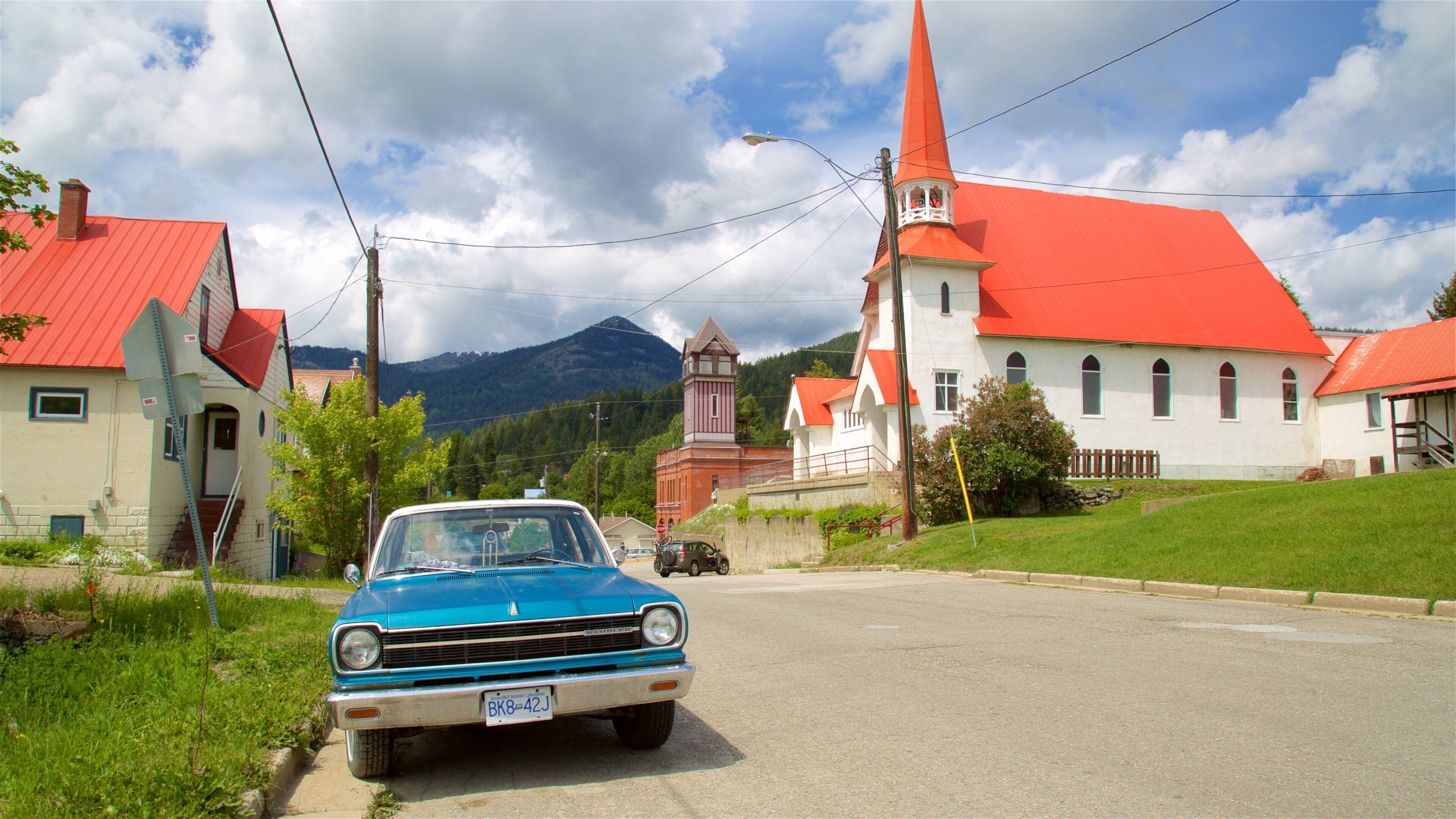 A blue vintage car parked on a rural road, a church with a red roof, and a mountain in the background.