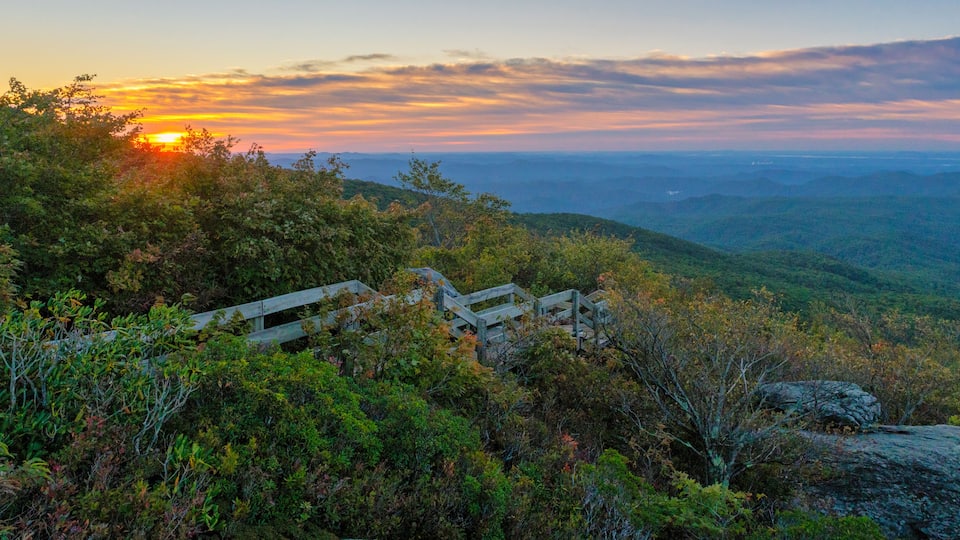 A sunset over a mountainous landscape with a wooden bridge and dense foliage.