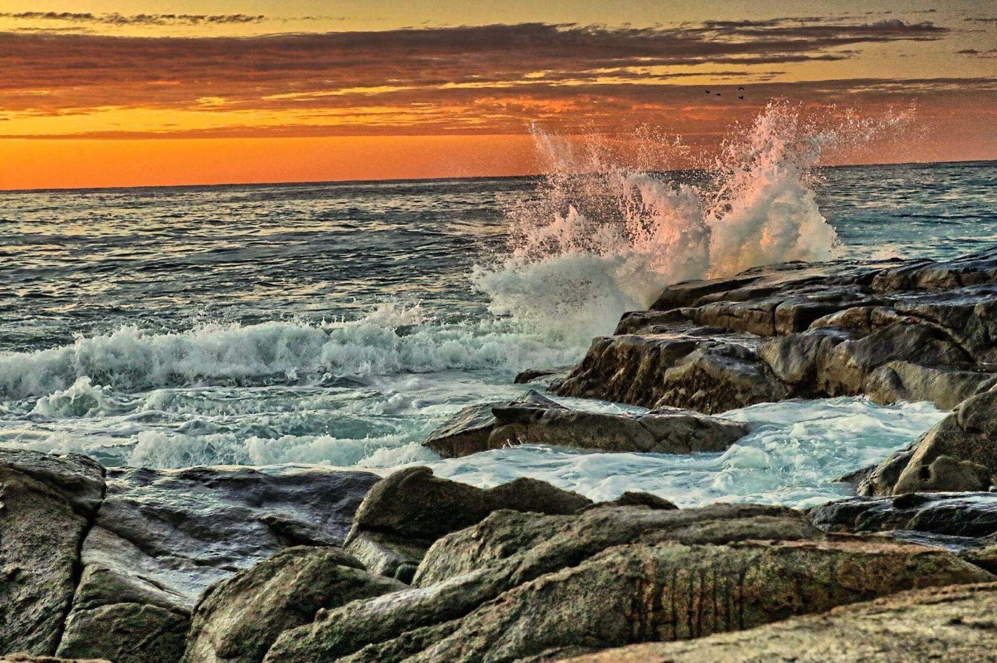 A rocky shoreline with waves crashing against the rocks during sunset.