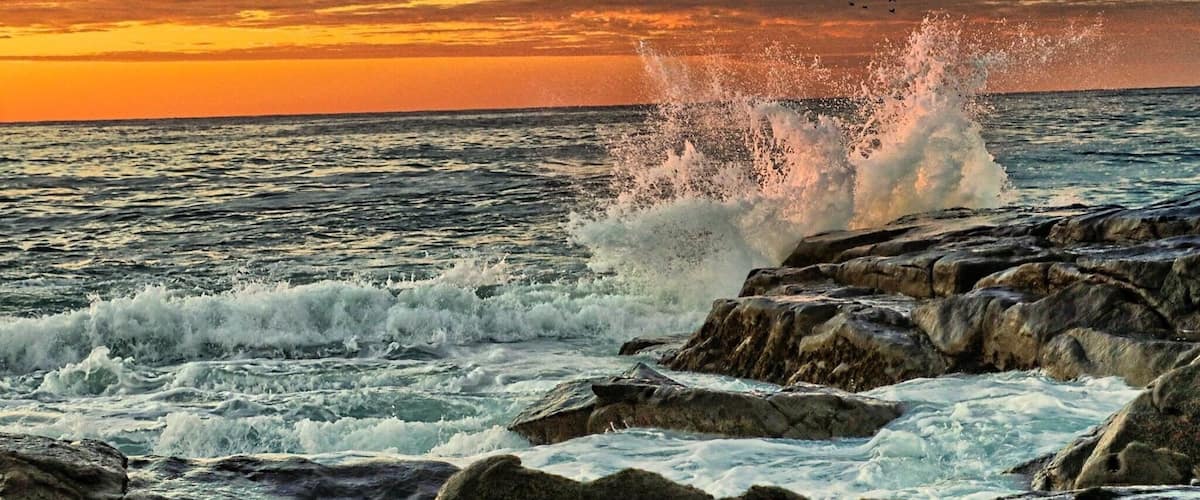 A rocky shoreline with waves crashing against the rocks during sunset.