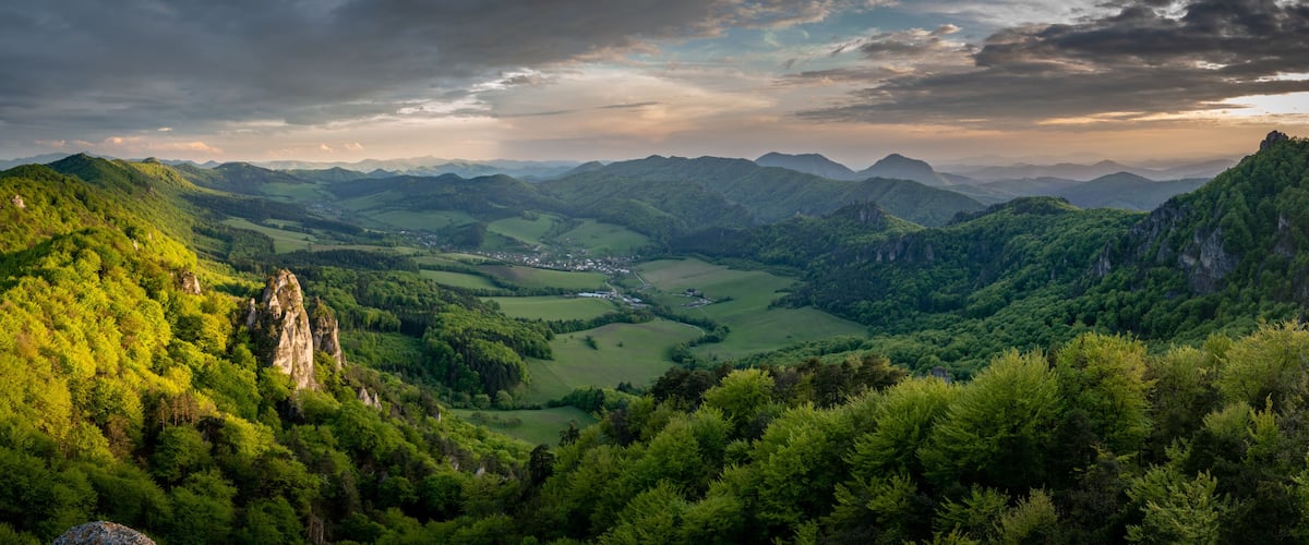A mountainous landscape with dense forests, a valley, and distant mountains under a cloudy sky.