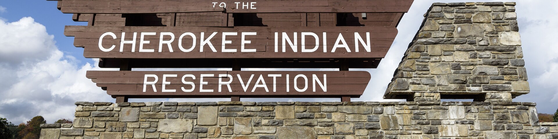 A wooden sign with 'Welcome to the Cherokee Indian Reservation' written on it, set against a stone wall and a clear blue sky.