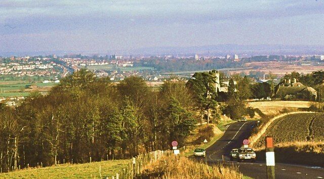 The A361 above Wroughton These days the road is numbered as A4361, for reasons better known to the highway authorities. The view down the hill includes the junction with the minor road to Elcombe, the tower of Wroughton parish church and the buildings of Swindon in the distance.