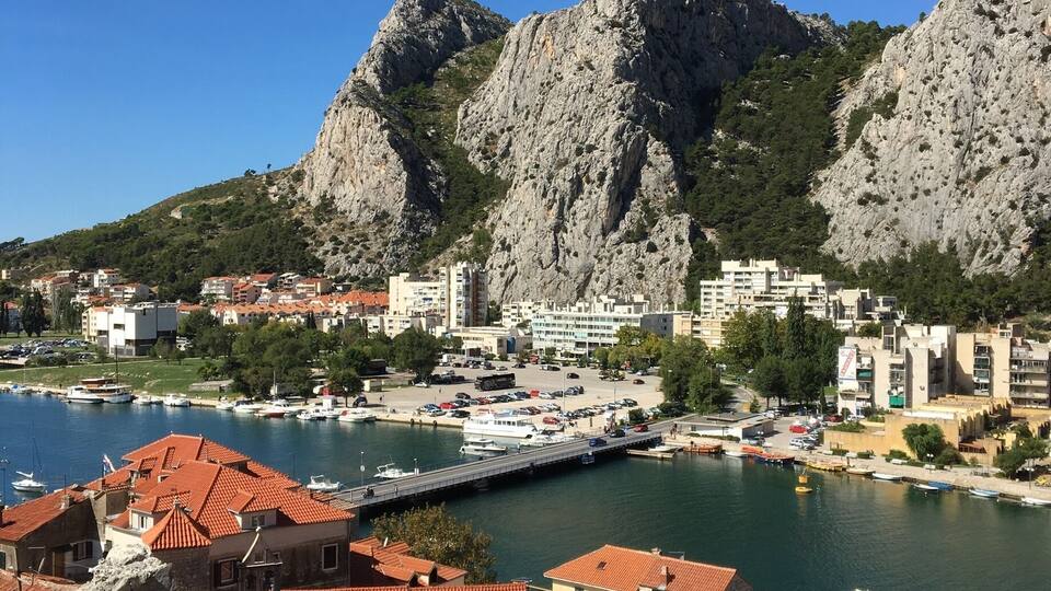 A coastal town with red-roofed buildings, a marina, and rocky mountains in the background.
