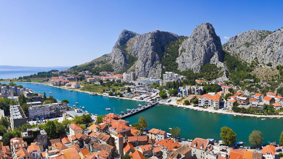 A coastal town with red-roofed buildings, a marina, and rocky mountains in the background.