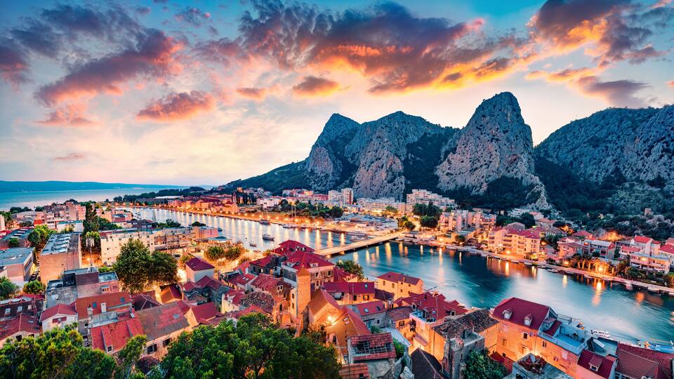 A coastal town with a river, red-roofed buildings, and mountains in the background.