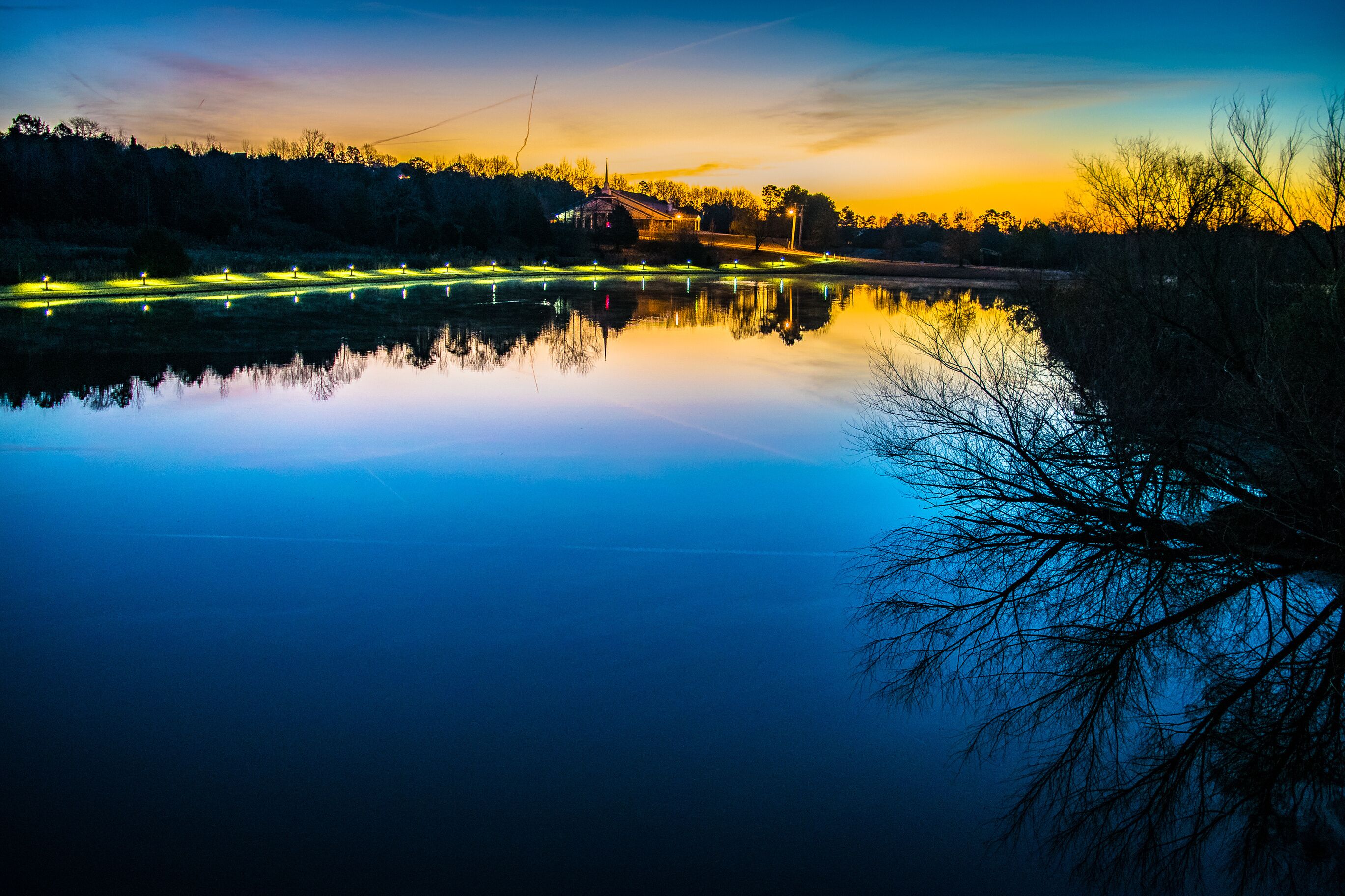A calm lake at dusk with reflections of trees and lights, surrounded by a forested area.