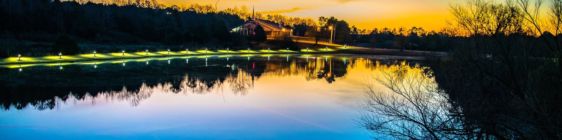 A calm lake at dusk with reflections of trees and lights, surrounded by a forested area.