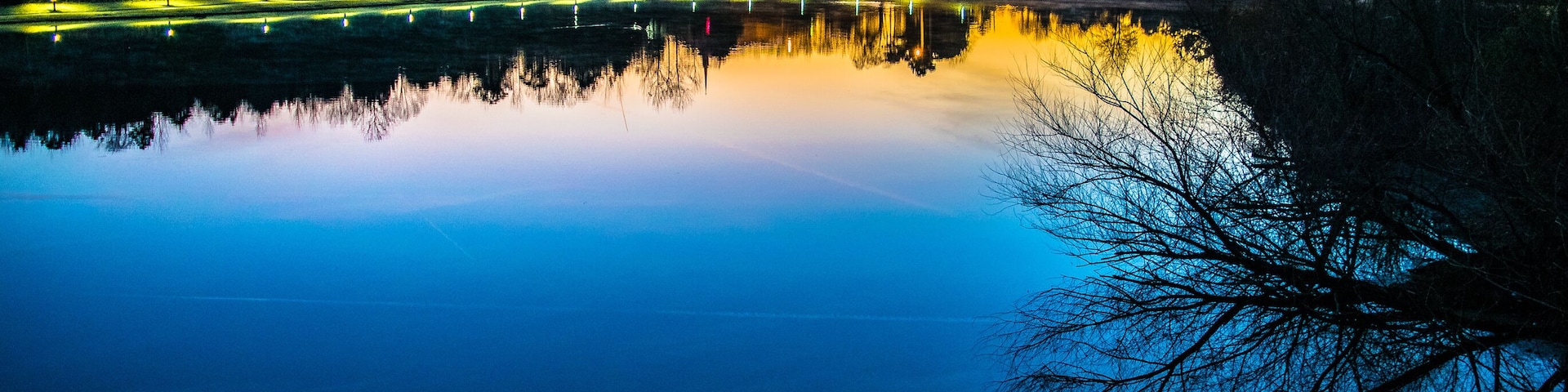 Um lago tranquilo ao entardecer, com reflexos de árvores e luzes, cercado por uma área arborizada.