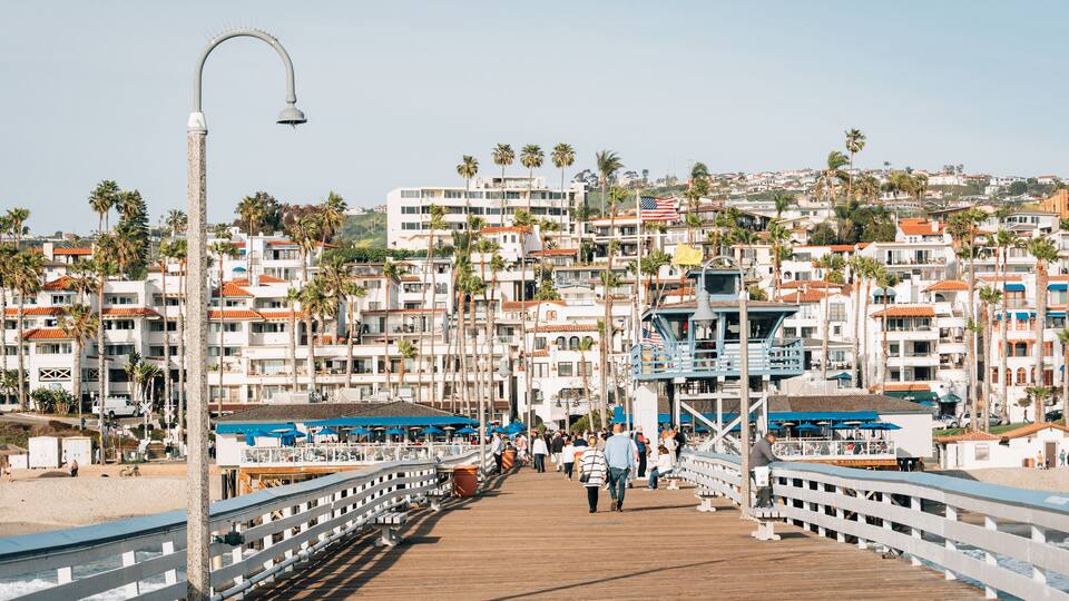 A boardwalk with people walking, palm trees, and buildings in the background.