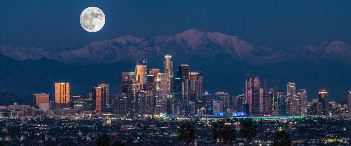 A city skyline with illuminated buildings and a full moon in the sky, with snow-capped mountains in the background.