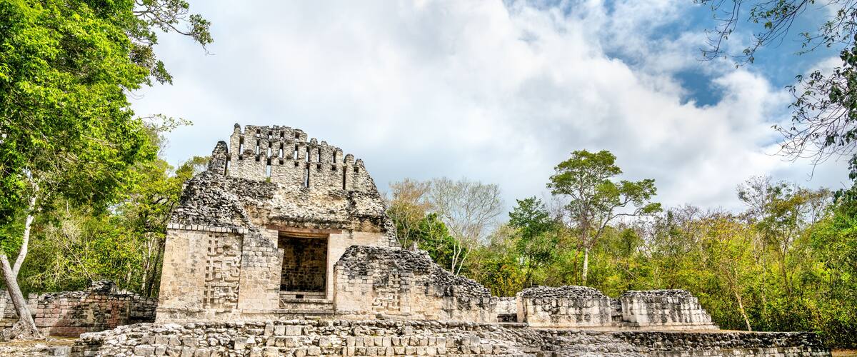 An ancient stone structure with a central doorway, surrounded by lush greenery and a partly cloudy sky.