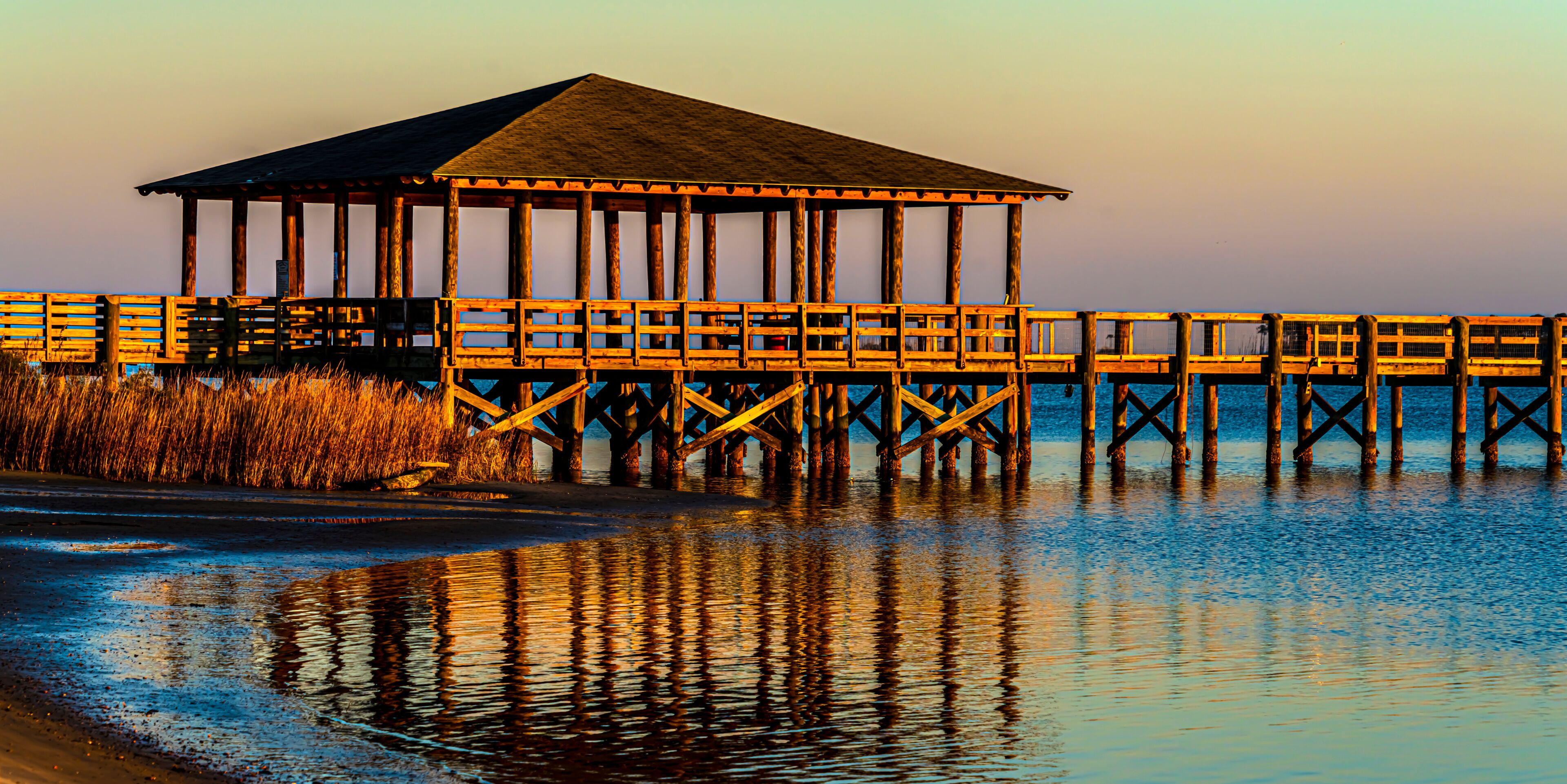 A wooden pier with a covered structure extending into calm waters, surrounded by tall grasses.