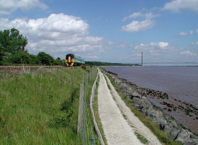 A train on tracks beside a river, a bridge in the distance, and a grassy path.