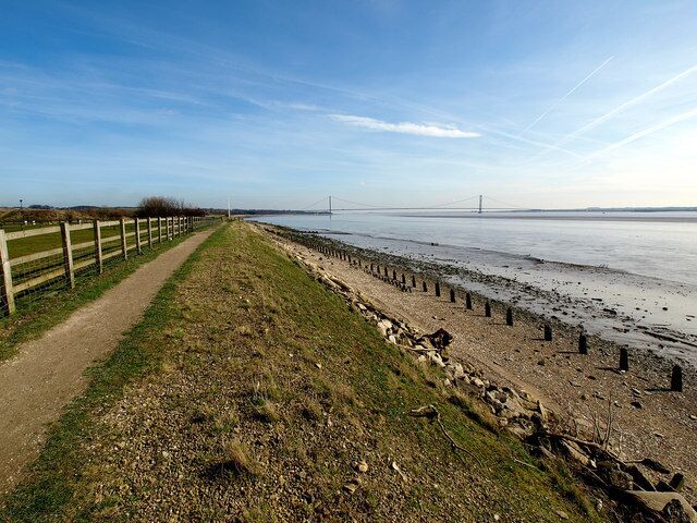 A coastal path with a fence, a sandy beach, and a bridge in the distance.