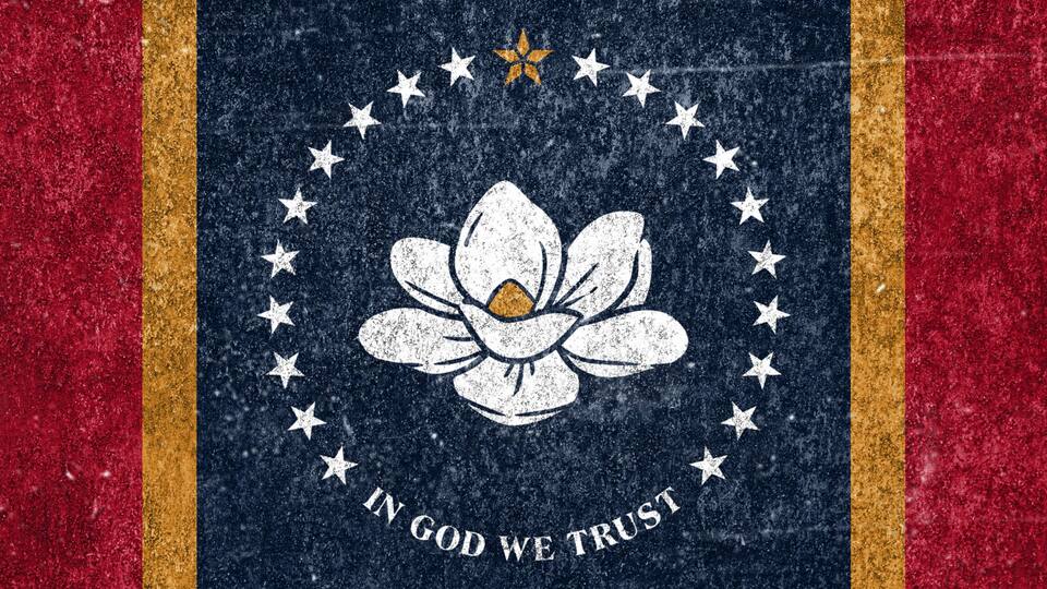 A flag with a white flower emblem, stars, and the phrase 'IN GOD WE TRUST' on a red background.