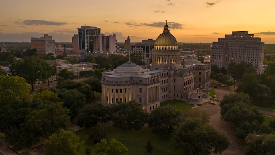 A large, historic building with a prominent dome, surrounded by modern skyscrapers and lush greenery.