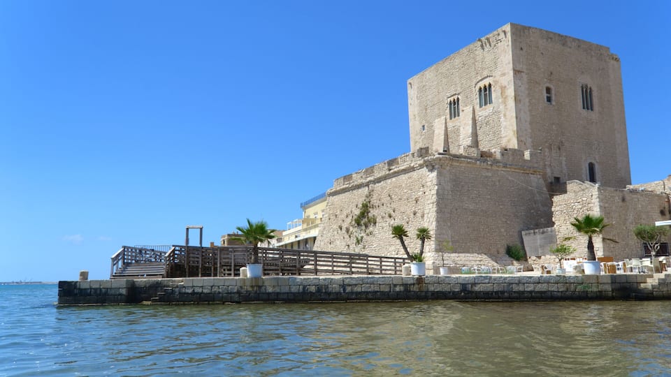 A historic stone building with a tower, situated by a calm body of water, with a wooden walkway leading to a viewing platform.