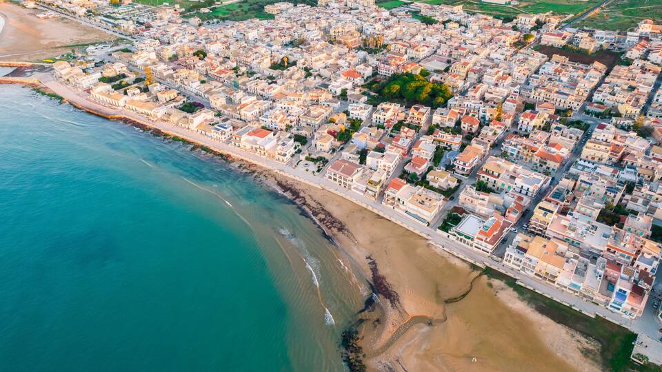 An aerial view of a coastal town with a beach, dense housing, and a clear blue sea.
