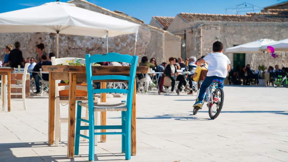 A turquoise chair in a plaza with tables and umbrellas, a person riding a bicycle, and stone buildings in the background.