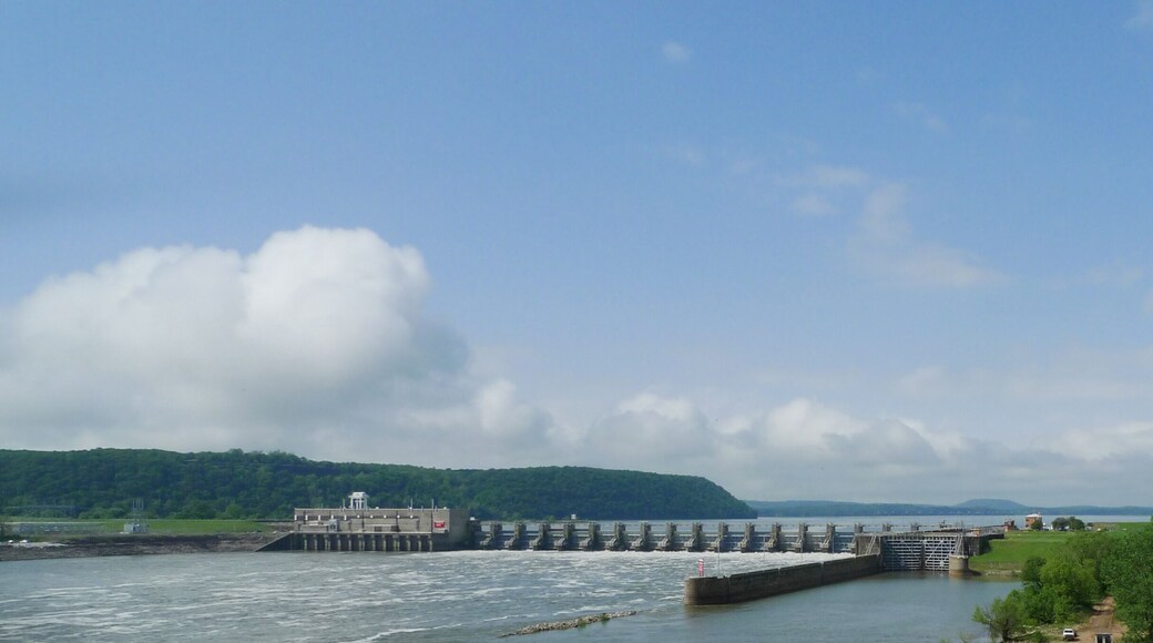 A dam with a lock system, a river, and a green landscape with trees and hills in the background.