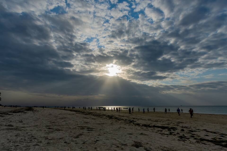 A beach with people walking, the sun shining through clouds, and the ocean in the background.