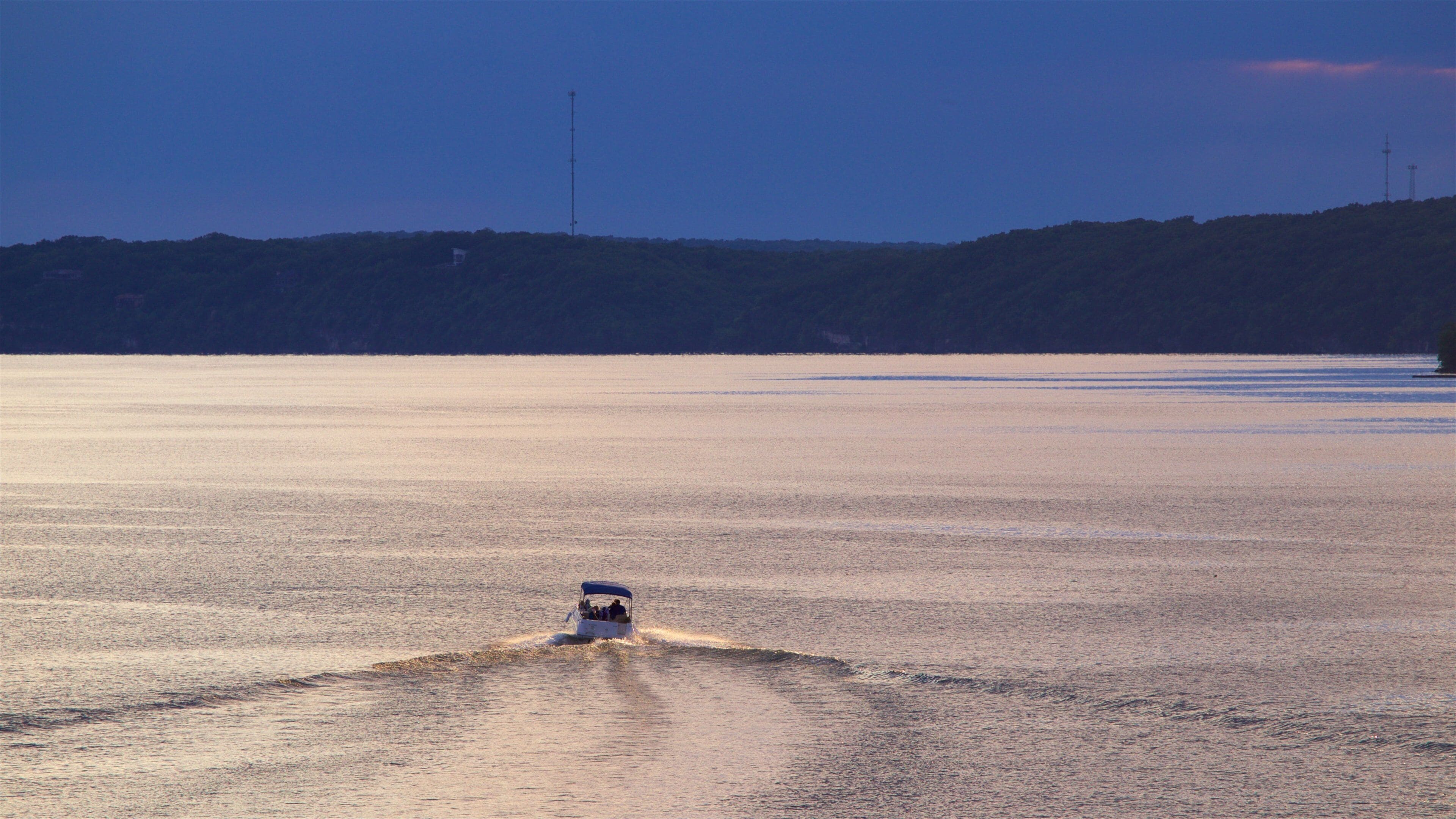 A boat creating a wake on a calm body of water with a hilly landscape in the background.