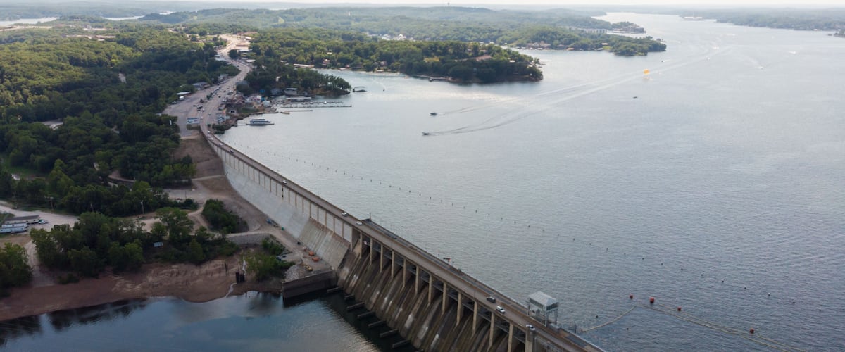 A large dam with a road running alongside it, a river, and a forested area in the background.