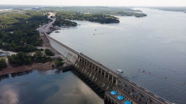 A large dam with a road running alongside it, a river, and a forested area in the background.
